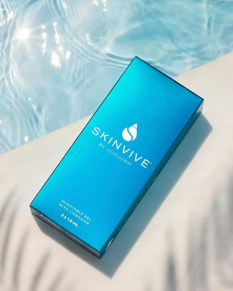 A single blue box of SKINVIVE by JUVÉDERM injectable gel, featuring a silver logo and product name, resting on a white, textured surface next to a sunlit pool with rippling water. The packaging specifies "INJECTABLE GEL WITH LIDOCAINE" and "2 x 1.0 mL.