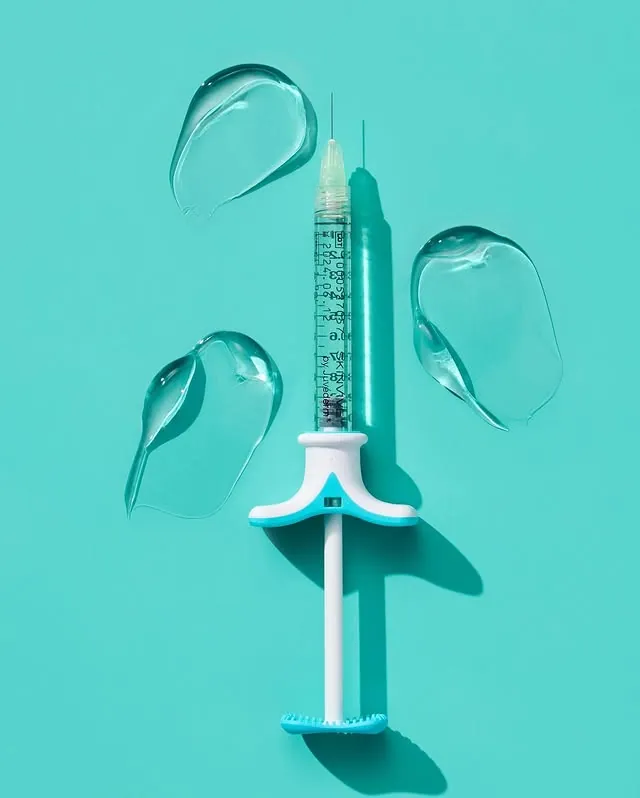 A close-up, top-down shot of a SKINVIVE by JUVÉDERM injectable gel syringe on a light blue background. The syringe is surrounded by four clear, gel-like swooshes, suggesting the product's texture and function for improving skin smoothness.